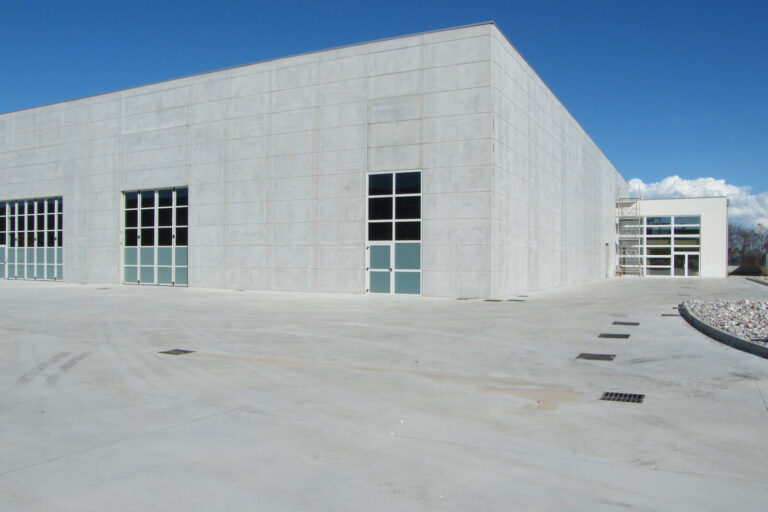 Finestre e Portoni Design per Edifici Commerciali: La Nostra Esperienza ad Affi (VR) Immagine esterna di un moderno capannone industriale o commerciale, caratterizzato da ampie pareti in cemento prefabbricato di colore chiaro. L'edificio ha un design pulito e lineare. In primo piano, su due facciate angolari, sono visibili grandi finestre a tutta altezza con infissi bianchi che formano una griglia, alcune delle quali sembrano essere delle porte-finestre. Le finestre riflettono il cielo azzurro e il paesaggio circostante. L'area esterna è pavimentata in cemento chiaro, con alcune griglie di scolo sul terreno e aree con ghiaia. Sullo sfondo si notano altri edifici industriali e un cielo blu con qualche nuvola bianca.