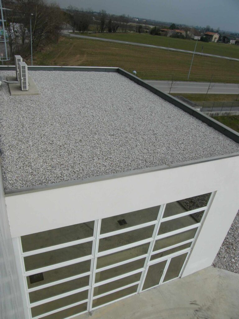 Design Funzionale: Serramenti per Capannoni e Edifici Commerciali ad Affi (VR) Immagine aerea o dall'alto di una parte di un moderno capannone industriale o edificio commerciale situato ad Affi (VR). L'edificio ha pareti esterne bianche e un ampio tetto piatto ricoperto di ghiaia chiara, sul quale sono visibili alcune unità esterne di condizionamento o ventilazione. La facciata in primo piano è caratterizzata da una grande superficie vetrata con infissi chiari che formano una griglia geometrica, riflettendo parzialmente il cielo e l'ambiente circostante. Intorno all'edificio si intravede un'area esterna con vegetazione sparsa, strade e altri edifici in lontananza, sotto un cielo leggermente nuvoloso.