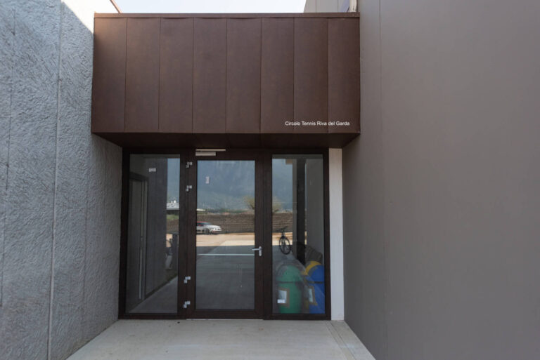 Porte a Vetro e Chiusure per Strutture Sportive: Circolo Tennis a Riva del Garda (TN) Immagine ravvicinata di un ingresso moderno in un edificio, identificato come "Circolo Tennis Riva del Garda", con pareti esterne di colore grigio e una sezione superiore rivestita in pannelli marroni. L'ingresso è costituito da una serie di porte a battente in vetro con infissi marroni scuri. Attraverso i vetri, si intravedono un parcheggio, un'auto e una bicicletta. Sulla parete marrone sopra le porte, è scritto "Circolo Tennis Riva del Garda".