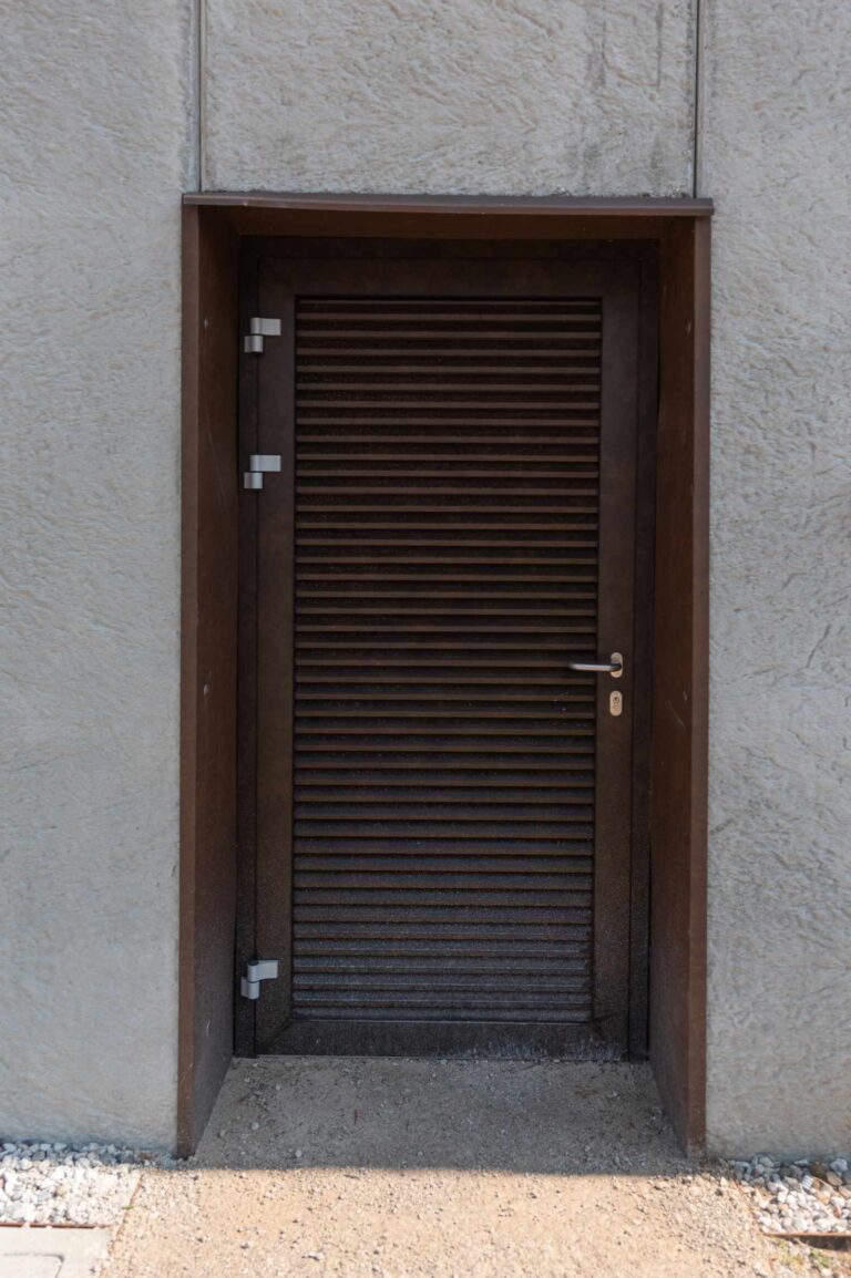 Design Industriale e Durevole: I Nostri Serramenti per il Circolo Tennis di Riva del Garda (TN) Immagine ravvicinata di una porta di servizio in metallo marrone scuro, con una superficie a lamelle orizzontali, installata in una parete di cemento grigio chiaro con texture ruvida. La porta è incorniciata da una struttura marrone scuro che la circonda su tre lati. In alto, sopra la porta, è visibile una piccola sporgenza orizzontale. La maniglia e la serratura della porta sono in metallo argentato. Il pavimento davanti alla porta è composto da ghiaia chiara.