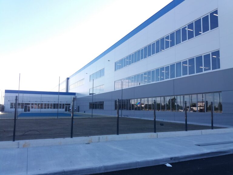 Architettura Funzionale per Grandi Spazi Commerciali: I Nostri Serramenti al Decathlon di Castel San Pietro Terme (BO) Immagine esterna di un grande edificio moderno, probabilmente il Decathlon a Castel San Pietro Terme (BO), con facciate bianche e grigie e un lungo nastro di finestre orizzontali in vetro con infissi bianchi. La parte superiore dell'edificio presenta una striscia orizzontale di colore blu. In primo piano, c'è un'area esterna con una recinzione metallica e un'area verde con erba secca. Sullo sfondo, si intravede un cielo azzurro e sereno.