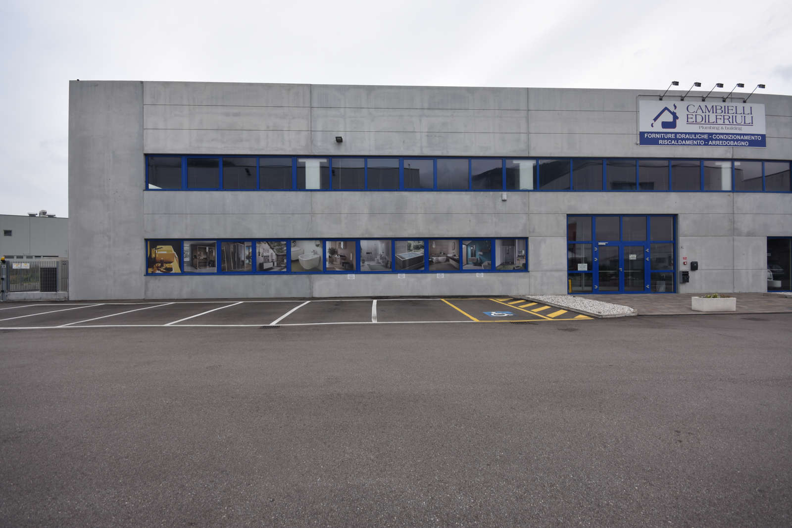 Progetto Edilizio Commerciale: Serramenti su Misura per Cambielli Edilfriuli a Gardolo (TN) Immagine esterna di un moderno capannone industriale o commerciale con facciata in pannelli di cemento prefabbricato di colore grigio chiaro. L'edificio presenta una lunga fila di finestre orizzontali con infissi blu scuro lungo il piano superiore, e una serie di finestre simili al piano inferiore, alcune delle quali mostrano immagini espositive di prodotti da bagno o arredi interni. Sulla destra della facciata, un ingresso principale con porte vetrate con infissi blu scuro. In alto a destra, è visibile un'insegna verticale luminosa con il logo "CAMBIELLI EDILFRIULI" e dettagli sui servizi offerti. L'area antistante l'edificio è un ampio parcheggio asfaltato con segnaletica orizzontale, inclusi posti per disabili. Il cielo è grigio e nuvoloso.