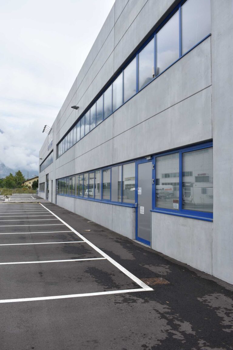 Realizzazione Serramenti Commerciali: Capannone con Facciata Moderna a Gardolo (TN) Immagine esterna di un moderno capannone industriale o commerciale, ripreso da un angolo che mostra due delle sue facciate. L'edificio è rivestito con pannelli di cemento prefabbricato di colore grigio chiaro e presenta lunghe finestre orizzontali con infissi blu scuro, che percorrono l'intera lunghezza delle facciate. Le finestre riflettono il cielo grigio e nuvoloso e parti del paesaggio circostante. In primo piano, un ampio parcheggio asfaltato con segnaletica orizzontale bianca per i posti auto. In lontananza, si intravedono alberi e montagne sotto un cielo coperto. L'immagine trasmette un senso di modernità e pulizia strutturale.