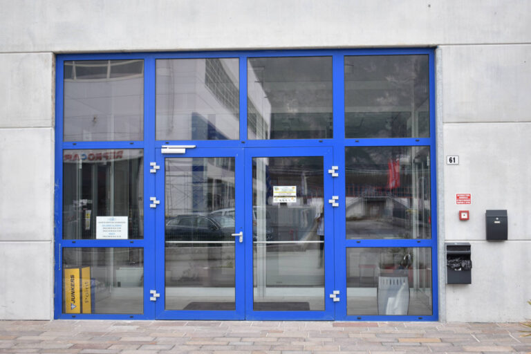 Porte e Finestre Industriali: Progetto Commerciale a Gardolo (TN) Un'immagine frontale di un ingresso moderno in un edificio commerciale o industriale. L'ingresso è costituito da un'ampia facciata vetrata con infissi di colore blu brillante, suddivisa in più pannelli rettangolari. Due di questi pannelli fungono da porte, con maniglie in metallo e serrature. Attraverso i vetri, si intravedono gli interni luminosi dell'edificio e, in lontananza, il riflesso di altri edifici e impalcature. Sulla destra dell'ingresso, sulla parete esterna di colore chiaro, sono visibili un numero civico "61", un estintore a muro rosso e una cassetta nera. Sul lato sinistro, è presente una colonna gialla con scritte e dei contenitori per la raccolta differenziata. Il pavimento esterno è una pavimentazione a blocchi di colore chiaro.