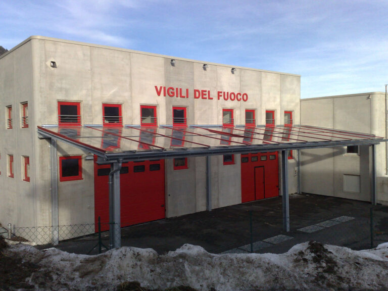 Portoni e Finestre Rosse: I Nostri Serramenti per la Caserma dei Vigili del Fuoco a Bleggio Superiore (TN) Ampia veduta frontale di una moderna caserma dei Vigili del Fuoco a Bleggio Superiore (TN), con facciate in cemento chiaro. L'edificio presenta una serie di finestre rettangolari e grandi portoni a saracinesca, tutti di un vivace colore rosso. Davanti all'ingresso principale, c'è una tettoia trasparente con struttura metallica che offre riparo. In primo piano, il terreno è parzialmente coperto di neve, suggerendo un clima freddo. Sullo sfondo, si intravedono montagne sotto un cielo azzurro con nuvole sparse.