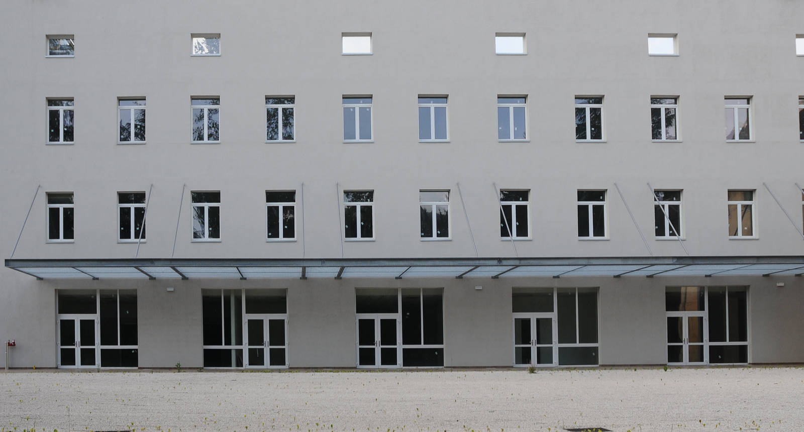 Chiusure e Vetrate per Edifici Pubblici: Sede Croce Rossa a Levico Terme (TN) Ampia veduta frontale di un edificio moderno a più piani, probabilmente la sede della Croce Rossa a Levico (TN), con una facciata chiara e uniforme. L'edificio presenta numerose finestre rettangolari con infissi bianchi, disposte simmetricamente su più livelli. Al piano terra, c'è una serie di ingressi vetrati con infissi bianchi, protetti da una lunga pensilina in vetro. Davanti all'edificio, si estende un'ampia area pianeggiante con ghiaia chiara, dove si notano piccole piante e una cassetta antincendio rossa a sinistra.
