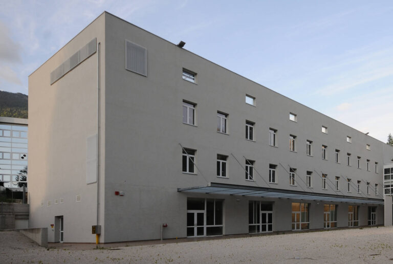 Finestre e Chiusure Innovative per la Sede della Croce Rossa a Levico Terme (TN) Veduta laterale di un edificio moderno a più piani, probabilmente la sede della Croce Rossa a Levico (TN), con facciate di colore grigio chiaro. L'edificio presenta numerose finestre rettangolari con infissi bianchi, disposte in modo irregolare. Al piano terra, c'è una lunga pensilina in vetro che copre una serie di ingressi vetrati. Sul lato sinistro dell'edificio, sono visibili delle griglie di ventilazione e un ingresso secondario. Davanti all'edificio, un'area esterna con ghiaia chiara. Sullo sfondo, si stagliano le montagne sotto un cielo chiaro.