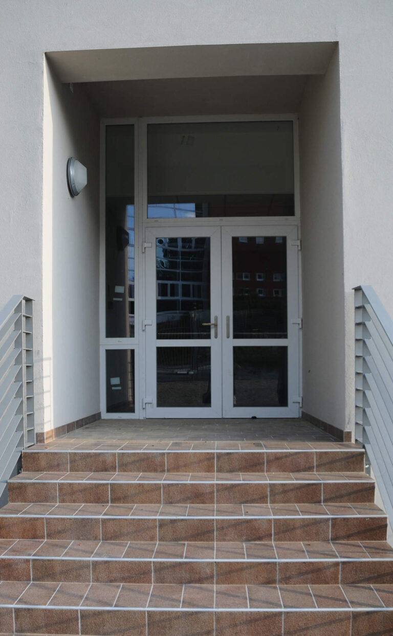 Porte a Vetro e Chiusure per la Sede della Croce Rossa a Levico Terme (TN) Immagine ravvicinata di un ingresso moderno in un edificio, probabilmente la sede della Croce Rossa a Levico (TN). L'ingresso è composto da una doppia porta a vetri con infissi bianchi, sormontata da una finestra orizzontale, anch'essa con infissi bianchi. Le porte si aprono su una scala di quattro gradini rivestiti in piastrelle marroni, con ringhiere metalliche sui lati. La parete circostante è di colore bianco, con una luce rotonda installata a sinistra dell'ingresso.