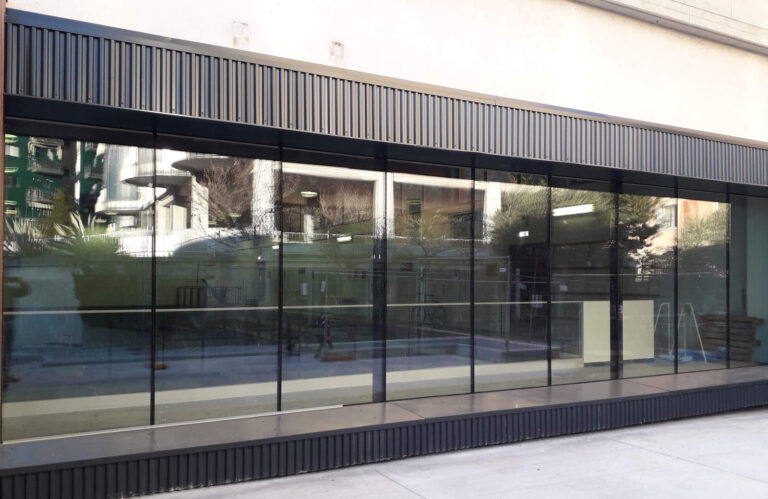 Design Contemporaneo e Luminosità: Le Nostre Chiusure per la Palestra Barattieri di Rovereto (TN) Veduta esterna di una moderna facciata della palestra Barattieri a Rovereto (TN). La facciata è dominata da una lunga e ampia vetrata che riflette l'ambiente circostante. Sopra e sotto le vetrate, ci sono pannelli scuri con texture a righe verticali. L'edificio è di colore chiaro con pilastri bianchi visibili. Il pavimento esterno è in cemento chiaro.