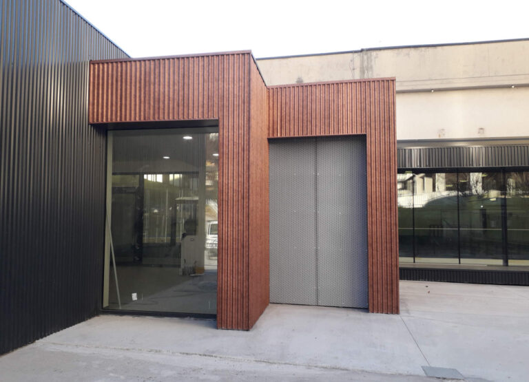 Facciate Innovativa e Ingressi Accoglienti per Centri Sportivi a Rovereto: Palestra Baratieri (TN) Veduta esterna di una parte della palestra Baratieri a Rovereto (TN), caratterizzata da una facciata moderna con pannelli verticali in legno scuro e ampie vetrate. Un ingresso con una porta a griglia metallica è incassato nella struttura. Il pavimento davanti all'edificio è in cemento chiaro. Sulla sinistra, si intravede una parete nera a doghe orizzontali, mentre sulla destra si notano altri elementi architettonici dell'edificio con vetrate che riflettono l'ambiente circostante.