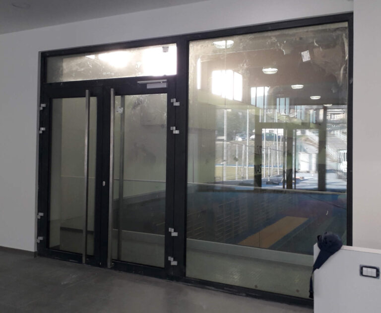 Realizzazione Serramenti Palestra Baratieri Rovereto (TN): Vetrate Luminose e Ingressi Moderni per Strutture Sportive Immagine interna di un ampio ambiente, probabilmente l'ingresso o una zona di transito della palestra Baratieri a Rovereto (TN). La scena è dominata da una grande vetrata con infissi neri e due porte a battente in vetro, che riflettono l'esterno e lasciano intravedere altre aree della palestra, inclusi campi da gioco e attrezzature sportive. La luce del sole filtra attraverso le finestre, creando riflessi sul vetro. Il pavimento è di colore grigio scuro, e una parete bianca è visibile sulla destra.