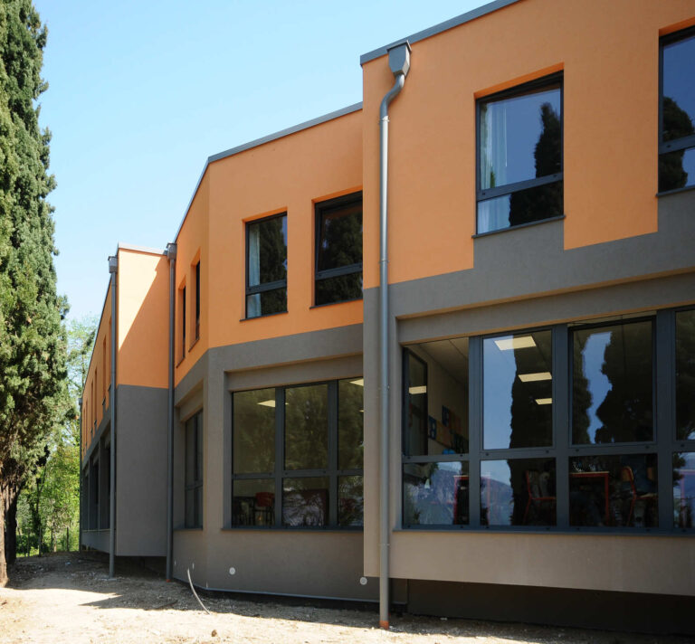 Innovazione e Luminosità per l’Apprendimento: Serramenti su Misura per il Liceo Sportivo Sacra Famiglia (VR) Veduta esterna di un moderno edificio scolastico a più piani, il Liceo Sportivo Sacra Famiglia a Castelletto di Brenzone (VR). La facciata è caratterizzata da un design a blocchi di colore, con sezioni arancioni chiare e grigio scure. Il piano terra presenta ampie vetrate con infissi scuri, che riflettono l'ambiente circostante. I piani superiori hanno finestre rettangolari con infissi scuri. L'edificio è affiancato da alti alberi di cipresso sul lato sinistro, e il terreno in primo piano è una combinazione di terra e ghiaia. Il cielo è sereno e azzurro.