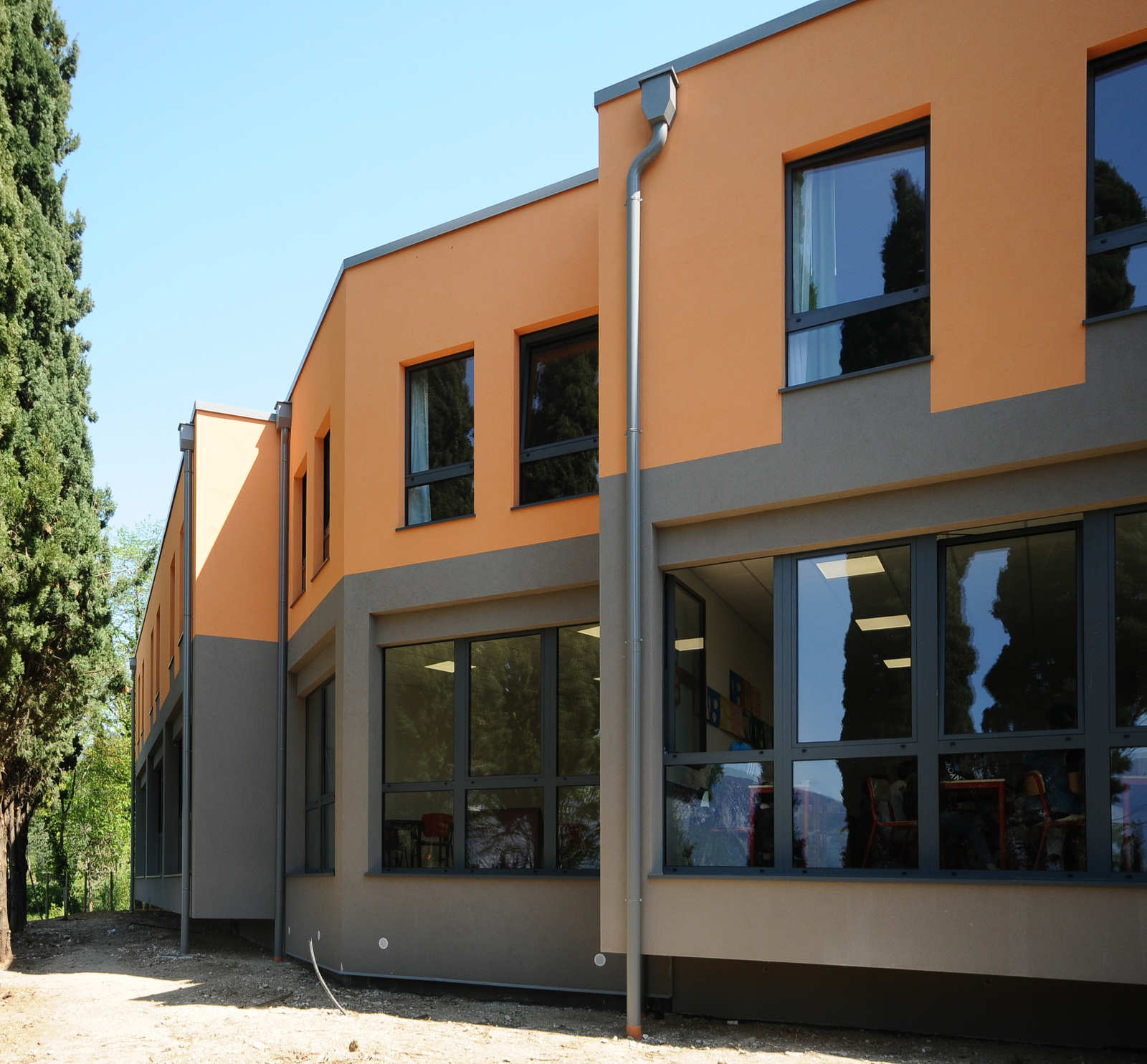 Innovazione e Luminosità per l’Apprendimento: Serramenti su Misura per il Liceo Sportivo Sacra Famiglia (VR) Veduta esterna di un moderno edificio scolastico a più piani, il Liceo Sportivo Sacra Famiglia a Castelletto di Brenzone (VR). La facciata è caratterizzata da un design a blocchi di colore, con sezioni arancioni chiare e grigio scure. Il piano terra presenta ampie vetrate con infissi scuri, che riflettono l'ambiente circostante. I piani superiori hanno finestre rettangolari con infissi scuri. L'edificio è affiancato da alti alberi di cipresso sul lato sinistro, e il terreno in primo piano è una combinazione di terra e ghiaia. Il cielo è sereno e azzurro.