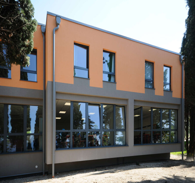 Spazi Funzionali e Accoglienti: Serramenti su Misura per Scuole a Castelletto, Verona Veduta esterna di un moderno edificio scolastico a due piani, il Liceo Sportivo Sacra Famiglia a Castelletto di Brenzone (VR). La facciata è distintiva, con la parte superiore di colore arancione chiaro e la parte inferiore grigio scuro. Il piano terra presenta ampie vetrate continue che riflettono l'ambiente esterno, mentre il piano superiore ha finestre più piccole con infissi scuri. Sul lato sinistro, è visibile una grondaia scura che scende lungo la facciata. Alcuni alberi di cipresso affiancano l'edificio sui lati.