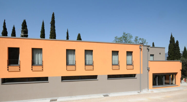 Innovazione e Qualità per la Didattica: I Nostri Serramenti al Liceo Sportivo Sacra Famiglia di Brenzone (VR) Veduta esterna di un moderno edificio scolastico a due piani, il Liceo Sportivo Sacra Famiglia a Castelletto di Brenzone (VR). La facciata principale è divisa orizzontalmente in due sezioni: la parte superiore è di colore arancione chiaro e presenta finestre rettangolari con piccole ringhiere, mentre la parte inferiore è grigio scuro e ha finestre orizzontali allungate. Sulla destra, un volume aggettante dell'edificio è anch'esso arancione chiaro e grigio scuro, con ampie vetrate. Dietro l'edificio, si ergono alberi di cipresso. Il cielo è azzurro e sereno.