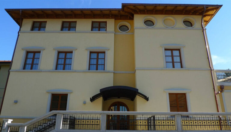 Design Innovativo e Sicuro: Le Nostre Chiusure per la Scuola Media di Mattarello (TN) Veduta frontale di un edificio moderno a più piani, le scuole medie di Mattarello (TN). La facciata è di colore giallo chiaro e presenta finestre rettangolari e, in una sezione verticale più stretta, finestre ovali. Il tetto è spiovente con tegole in terracotta e ampie gronde in legno. Un ingresso ad arco con una pensilina scura è visibile al piano terra, preceduto da una ringhiera decorata in ferro battuto.