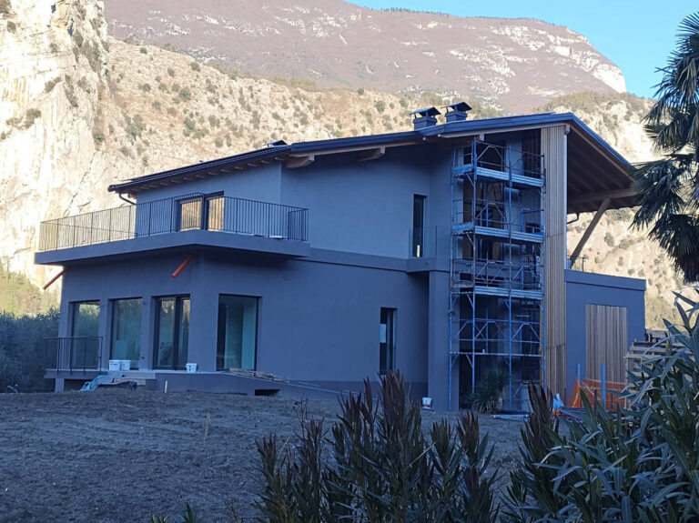 Realizzazione Serramenti Design ad Arco (TN): Villa Moderna in Costruzione Immagine di una grande villa moderna in fase avanzata di costruzione o ristrutturazione, situata ai piedi di montagne rocciose ad Arco (TN). L'edificio ha pareti esterne di colore grigio scuro e un tetto spiovente. Sono visibili ampie finestre con infissi scuri, alcune delle quali sembrano essere in fase di installazione o completamento. Sulla sinistra, c'è un balcone con una ringhiera orizzontale scura. La facciata frontale destra della casa presenta un'impalcatura che copre una sezione dove è visibile un rivestimento verticale in listelli di legno chiaro. Il terreno circostante è ancora in fase di sistemazione, con terra smossa in primo piano. Alcune piante e alberi sono visibili ai lati dell'inquadratura, con il paesaggio montano che domina lo sfondo.