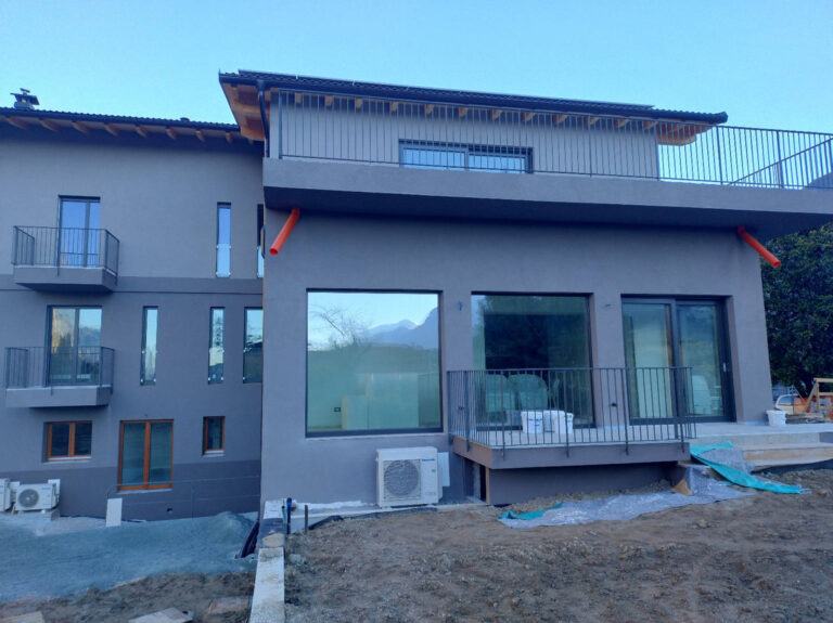 Serramenti e Chiusure Alta Qualità: Abitazione Privata ad Arco (TN) Completata Immagine esterna di una moderna abitazione privata ad Arco (TN), con un design architettonico contemporaneo e ampi serramenti. L'edificio è caratterizzato da facciate in intonaco grigio e tetti a falda con coperture scure. Si notano diverse finestre e porte-finestre con infissi scuri, alcune delle quali a tutta altezza, che riflettono parzialmente il paesaggio circostante. Ci sono balconi con ringhiere metalliche orizzontali scure. Sul lato destro, in primo piano, il terreno è ancora in fase di sistemazione, con terriccio e materiali da cantiere. Sotto un'ampia finestra al piano terra, è visibile un'unità esterna di un condizionatore. Il cielo è limpido e sullo sfondo si intravedono le montagne della zona.