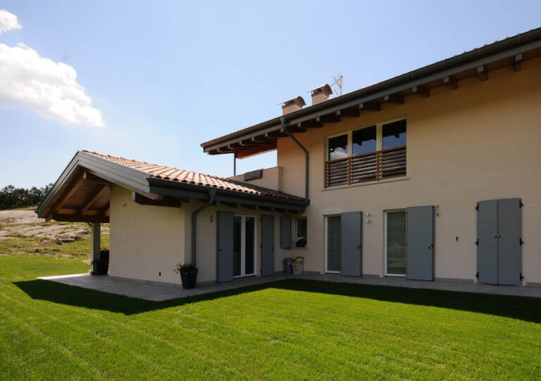 Finestre e Scuri Design: Realizzazione per Ville Moderne a Toldi (TN) Immagine esterna di una spaziosa abitazione privata di architettura moderna, situata in un contesto rurale e soleggiato. La casa ha facciate di colore beige chiaro e un tetto tradizionale con tegole rosse e ampie gronde in legno a vista. Si notano diverse aperture: ampie porte-finestre e finestre con infissi chiari, alcune delle quali sono dotate di scuri esterni a lamelle di colore grigio chiaro, parzialmente aperti. Al piano superiore, una finestra ha una ringhiera decorativa in legno. La casa è circondata da un prato verde ben curato che si estende in primo piano. Sullo sfondo si intravede la vegetazione e un terreno collinare, con un cielo azzurro e alcune nuvole.