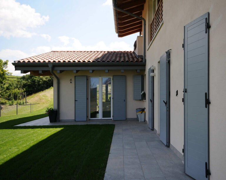 Serramenti e Scuri Design: Realizzazione per Residenza Privata a Toldi, Trentino Immagine esterna di una moderna abitazione privata con facciate di colore chiaro e tetto in tegole rosse, situata in un contesto verde. In primo piano, un'ampia area con prato verde ben curato e un vialetto pavimentato in piastrelle grigie che costeggia la casa. Sono visibili diverse porte-finestre e finestre dotate di scuri esterni a lamelle di colore grigio chiaro, alcuni dei quali aperti e altri chiusi. La casa presenta anche un portico con travi in legno a vista. Sullo sfondo, si intravede la vegetazione circostante, suggerendo una posizione tranquilla e immersa nella natura.
