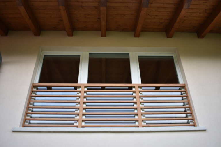 Design Moderno: Frangisole in Legno per Finestre di Case Privata a Toldi (TN) Immagine ravvicinata di una finestra moderna con un design distintivo, incastonata in una parete di colore chiaro. La finestra è composta da tre vetri con infissi bianchi. Sotto i vetri, si trova un sistema di frangisole orizzontale in legno chiaro, con lamelle fisse, che funge anche da parapetto decorativo. Sopra la finestra, è visibile una parte del tetto con travi in legno a vista, tipiche di una struttura moderna o tradizionale rivisitata. Il cielo è parzialmente riflesso nei vetri della finestra.