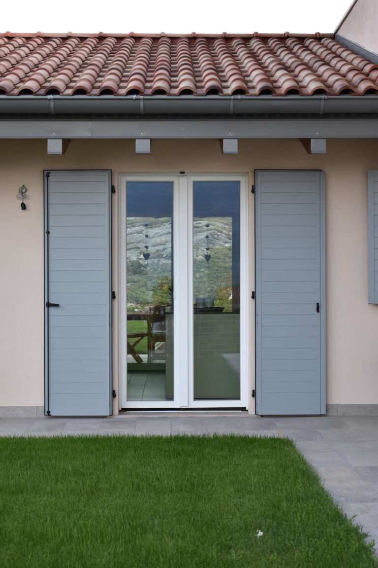 Porta Finestra e Scuri Design a Toldi (TN): Realizzazione Serramenti Esterni Immagine ravvicinata di una porta-finestra bianca con doppi vetri, affiancata da due scuri esterni a lamelle di colore grigio chiaro, parzialmente aperti. La porta-finestra riflette il paesaggio montano circostante. Sopra la finestra si nota un tetto tradizionale con tegole rosse e un sistema di grondaia. La parete esterna è di colore chiaro. Sotto la porta-finestra, c'è un piccolo patio pavimentato in piastrelle grigie che si affaccia su un prato verde curato. A sinistra, sulla parete, è appeso un piccolo elemento decorativo. Siamo a Toldi (TN) vicino Rovereto.