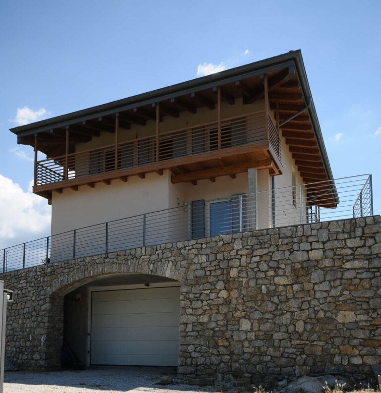 Architettura Contemporanea e Balconi Panoramici: I Nostri Serramenti a Toldi (TN) Immagine esterna di una moderna abitazione privata su più livelli, con una base imponente in pietra naturale a vista e una sovrastruttura con pareti di colore chiaro e un tetto a falda con ampie gronde in legno e travi a vista. Il piano terra presenta un arco in pietra che funge da ingresso a un garage con una porta bianca. Sopra la base in pietra, la casa è dotata di ampi serramenti orizzontali e verticali con infissi scuri. Un lungo balcone con ringhiere orizzontali in metallo corre lungo la facciata superiore. Un altro balcone con parapetto simile è visibile al piano intermedio. L'abitazione è immersa in un paesaggio collinare con cielo azzurro e alcune nuvole, suggerendo una posizione panoramica.