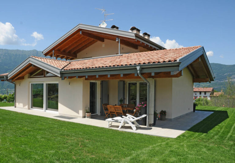 Realizzazione Serramenti Design a Toldi (TN): Abitazione Privata con Grandi Vetrate Immagine esterna di una spaziosa abitazione privata di recente costruzione o ristrutturazione, situata in una zona verde. La casa presenta facciate di colore chiaro e un tradizionale tetto in tegole rosse con ampie gronde in legno a vista. Sono visibili grandi porte-finestre e finestre con infissi chiari, alcune delle quali con scuri esterni chiusi o semi-aperti di colore grigio. Un patio esterno pavimentato ospita un tavolo e sedie in legno, oltre a due lettini prendisole bianchi e vasi di fiori colorati. L'abitazione è circondata da un prato verde ben curato e sullo sfondo si intravedono colline e montagne, tipiche del paesaggio trentino. Il cielo è azzurro con alcune nuvole sparse.