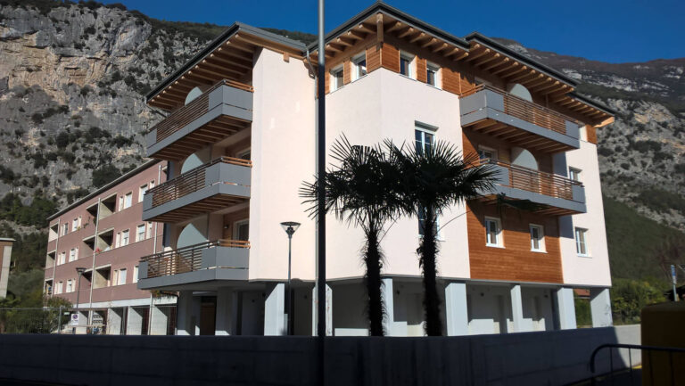 Finestre e Chiusure Alta Qualità per Edilizia Residenziale Pubblica a Dro (TN) Immagine esterna di un moderno complesso residenziale a più piani situato in un ambiente montano a Dro (TN). L'edificio presenta una combinazione di facciate in intonaco chiaro, pannelli di rivestimento in legno e balconi con parapetti orizzontali in metallo grigio scuro. Si notano numerose finestre con infissi bianchi e, in alcune sezioni, rivestimenti verticali in legno. La struttura del tetto è visibile con travi in legno a vista. Sullo sfondo si ergono imponenti pareti rocciose di montagna. In primo piano, due palme slanciate aggiungono un tocco di verde. L'edificio è ben illuminato dal sole, con un cielo azzurro e limpido.