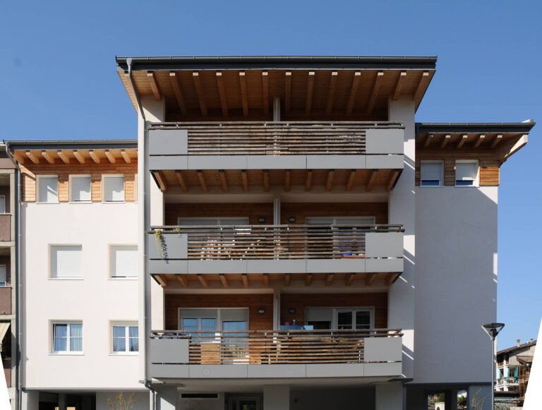 Realizzazione Serramenti Case ITEA Dro (TN): Balconi Moderni e Efficienza Energetica Immagine esterna frontale di un edificio residenziale a più piani, presumibilmente parte del complesso Case ITEA a Dro (TN). L'edificio è caratterizzato da facciate di colore chiaro e numerosi balconi a sbalzo con parapetti orizzontali in metallo grigio. Ogni balcone ha una sezione inferiore rivestita in legno chiaro a doghe orizzontali. Sono visibili diverse finestre con infissi bianchi e in alcuni casi scuri esterni chiari. La struttura del tetto è visibile con travi in legno a vista. L'edificio è ben illuminato dal sole, con un cielo azzurro e limpido. Sul lato sinistro si intravede un altro edificio simile.