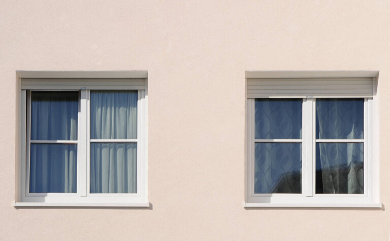 Realizzazione Serramenti Case ITEA Dro (TN): Finestre con Avvolgibili a Isolamento Termico Immagine ravvicinata di due finestre identiche con infissi bianchi su una facciata di colore chiaro, probabilmente parte di un edificio residenziale ITEA a Dro (TN). Entrambe le finestre hanno doppi vetri che riflettono parzialmente l'ambiente circostante. Sopra ciascuna finestra, è visibile un cassonetto per avvolgibili, uno dei quali è parzialmente abbassato, mostrando le lamelle. Le finestre sono incassate leggermente nella parete e presentano davanzali bianchi. La luce solare illumina uniformemente la facciata, mettendo in risalto la texture dell'intonaco.