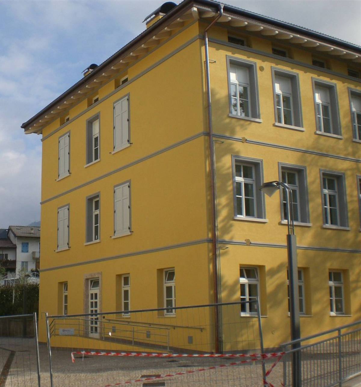 Riqualificazione Edilizia ITEA Miola (TN): I Nostri Serramenti su Facciate Gialle Immagine esterna di un moderno edificio residenziale a tre piani, probabilmente parte del complesso Case ITEA a Miola (TN), con facciate di un vivace colore giallo. L'edificio presenta una serie di finestre rettangolari con infissi grigio scuro e scuri esterni bianchi, alcuni aperti e altri chiusi. Il design include fasce orizzontali grigie che separano i piani. Sull'angolo dell'edificio è visibile una grondaia verticale metallica. In primo piano, l'area esterna è recintata con una bassa barriera metallica e nastro segnaletico rosso e bianco, suggerendo un cantiere o un'area di lavoro recente. Il cielo è azzurro con nuvole sparse.