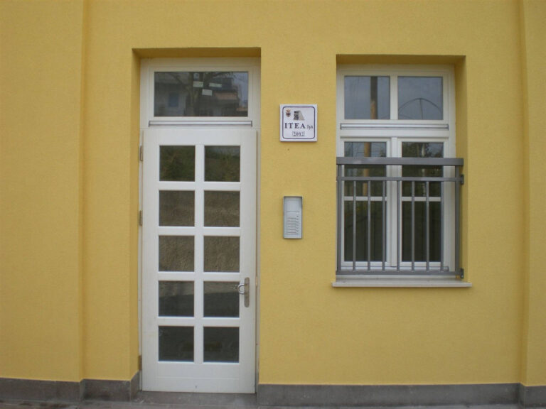 Porta d’Ingresso e Finestra con Ringhiera: Soluzioni Serramenti per Case ITEA Miola (TN) Immagine ravvicinata di una sezione della facciata gialla di un edificio residenziale, probabilmente parte delle Case ITEA a Miola (TN). La foto mostra una porta d'ingresso bianca a pannelli e una finestra adiacente, anch'essa con infissi bianchi. La finestra è dotata di una piccola ringhiera orizzontale in metallo scuro. Tra la porta e la finestra, sulla parete, sono installati un citofono e una placca informativa con la scritta "ITEA SpA" e l'anno "2012". La facciata presenta una texture intonacata, e la base dell'edificio è rifinita con un cordolo scuro.