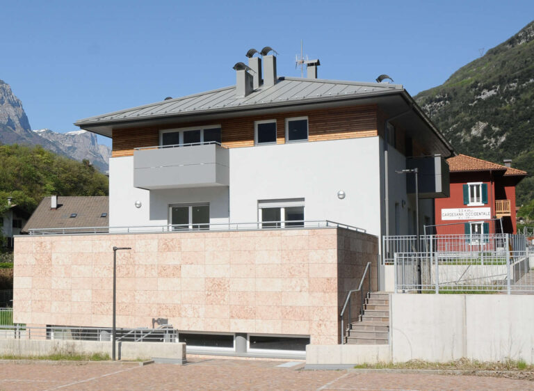 Finestre e Chiusure Alta Qualità per Complesso Residenziale ITEA a Padergnone, Trentino Immagine esterna di un moderno edificio residenziale a più piani, probabilmente parte del complesso Case ITEA a Padergnone (TN). L'edificio presenta una facciata principale di colore bianco con un rivestimento in pietra chiara al piano terra. Sono visibili ampie finestre rettangolari con infissi bianchi e balconi con parapetti grigi orizzontali. Il tetto è spiovente, di colore grigio, con travi in legno a vista e comignoli multipli. Sul lato destro, in secondo piano, si intravede una casa tradizionale rossa, e in lontananza, si ergono le montagne con vegetazione. In primo piano, c'è un'area pavimentata, probabilmente un parcheggio, con linee bianche di delimitazione. Il cielo è azzurro e limpido.