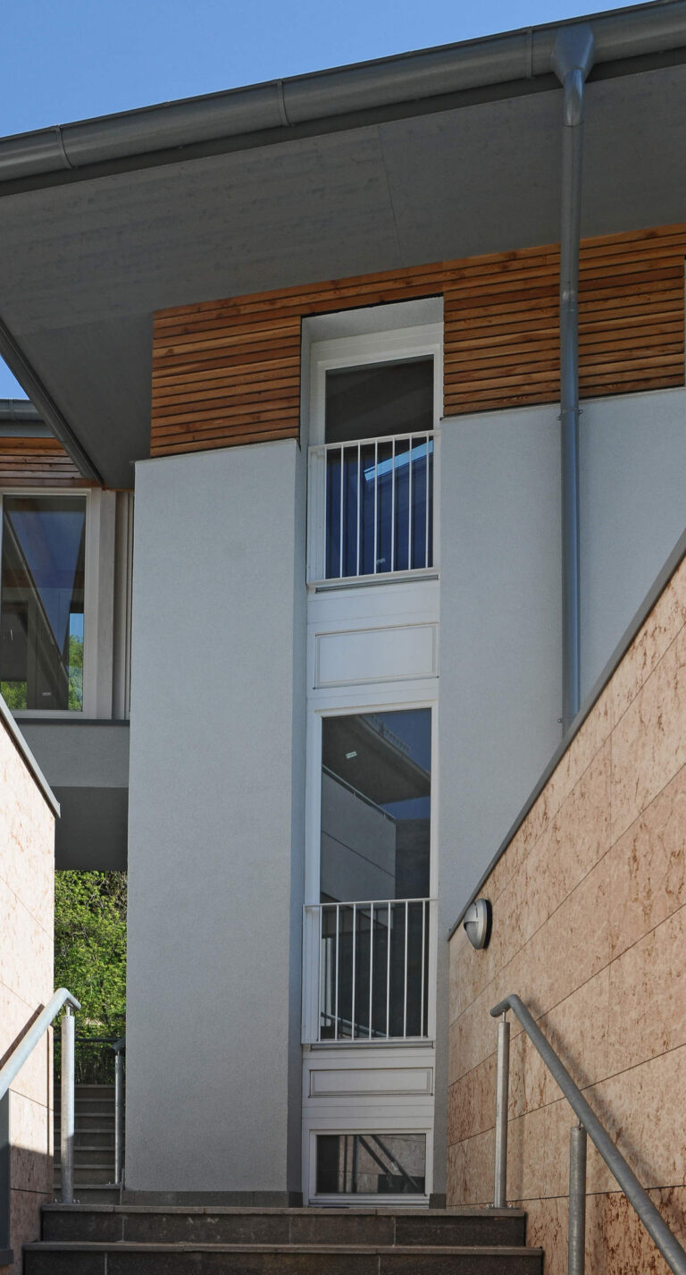 Finestre e Chiusure Alta Qualità per Complesso ITEA a Padergnone (TN) Immagine esterna verticale di una sezione di un moderno edificio residenziale, probabilmente parte del complesso Case ITEA a Padergnone (TN). La facciata è di colore chiaro con una sezione superiore rivestita in doghe di legno chiaro. Sono visibili tre finestre a tutta altezza, una sopra l'altra, con infissi bianchi. Le due finestre superiori sono dotate di piccole ringhiere metalliche orizzontali di colore bianco, mentre la finestra più in basso ha un davanzale a livello del terreno. Una grondaia metallica scende verticalmente sul lato destro della facciata. Sulla destra, una scala esterna con parapetti metallici e rivestimento in pietra conduce all'ingresso dell'edificio. Sullo sfondo, si intravede vegetazione lussureggiante e un cielo azzurro.