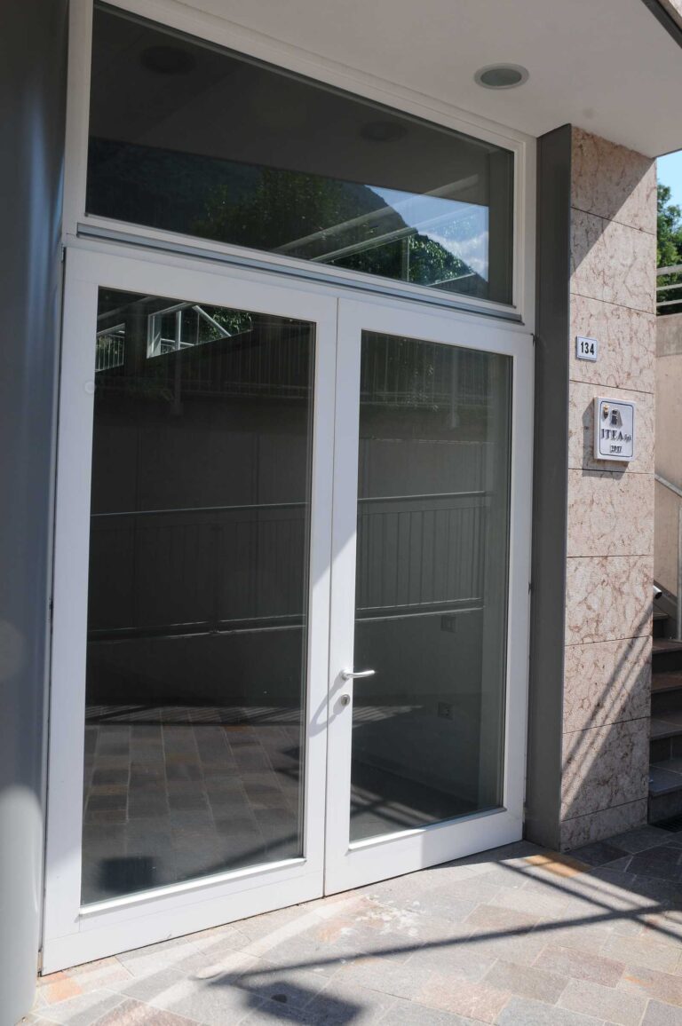 Porte a Vetro Alta Qualità per Complesso Residenziale ITEA a Padergnone (TN) Immagine esterna frontale di un ingresso moderno in un edificio residenziale, probabilmente parte del complesso Case ITEA a Padergnone (TN). L'ingresso è composto da una doppia porta a battente in vetro con infissi bianchi e un ampio pannello superiore fisso in vetro. I vetri riflettono il verde circostante e il cielo. La porta è affiancata da una colonna grigia a sinistra e una parete rivestita in pietra chiara sulla destra. Su quest'ultima è montato un numero civico "134" e una placca informativa con il logo "ITEA SpA" e l'anno "2012". Una scala in pietra sale lungo la parete destra. Il pavimento in primo piano è una pavimentazione esterna in piastrelle.