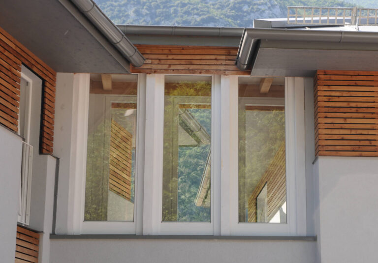 Finestre Triple e Chiusure Innovative per Complesso Residenziale ITEA a Padergnone (TN) Immagine ravvicinata di una sezione della facciata di un edificio moderno, probabilmente parte del complesso Case ITEA a Padergnone (TN). Si notano tre ampie finestre affiancate con infissi bianchi, che riflettono il paesaggio circostante, inclusi alberi e un cielo azzurro. Sopra e sotto le finestre, la facciata è rivestita con pannelli orizzontali in legno chiaro, aggiungendo un elemento caldo al design. Sopra la sezione in legno e la finestra, è visibile una grondaia metallica grigia. La parete è di colore chiaro, e in alto a destra si intravede la cima di una montagna.