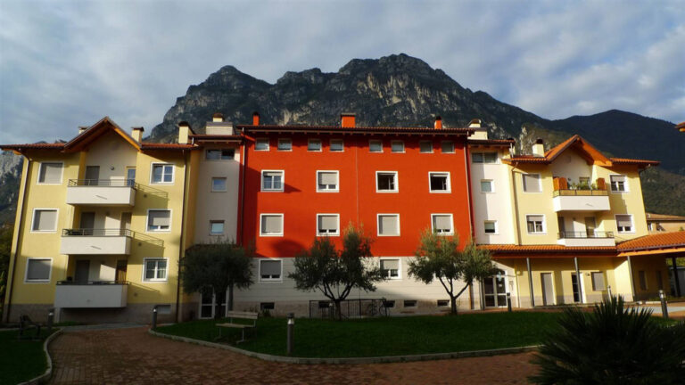 Architettura e Sviluppo Urbano: I Nostri Serramenti per Case ITEA a Riva del Garda (TN) Un'ampia veduta frontale di un complesso residenziale moderno con più edifici, situato a Riva del Garda (TN), sullo sfondo di imponenti montagne. L'edificio centrale è di un vivace colore rosso, mentre quelli laterali sono gialli e beige. Tutti gli edifici presentano numerose finestre quadrate o rettangolari con infissi bianchi e avvolgibili grigi. Alcuni edifici hanno balconi con parapetti in metallo o legno. In primo piano, c'è un'area pavimentata con mattoni e un prato verde ben curato, con alcuni alberi di olivo e panchine. Il cielo è nuvoloso ma luminoso.