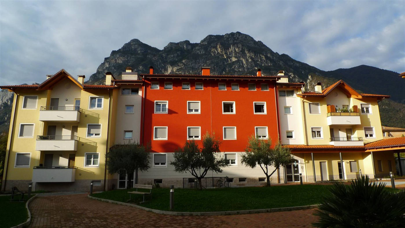 Architettura e Sviluppo Urbano: I Nostri Serramenti per Case ITEA a Riva del Garda (TN) Un'ampia veduta frontale di un complesso residenziale moderno con più edifici, situato a Riva del Garda (TN), sullo sfondo di imponenti montagne. L'edificio centrale è di un vivace colore rosso, mentre quelli laterali sono gialli e beige. Tutti gli edifici presentano numerose finestre quadrate o rettangolari con infissi bianchi e avvolgibili grigi. Alcuni edifici hanno balconi con parapetti in metallo o legno. In primo piano, c'è un'area pavimentata con mattoni e un prato verde ben curato, con alcuni alberi di olivo e panchine. Il cielo è nuvoloso ma luminoso.