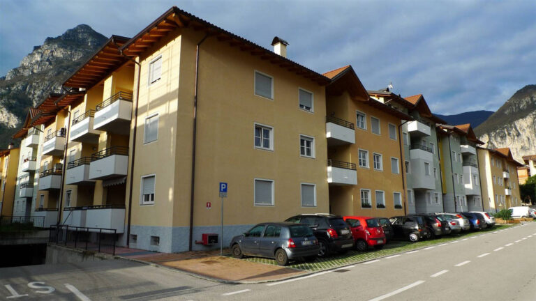 Finestre e Chiusure Alta Qualità per Complesso Residenziale ITEA a Riva del Garda (TN) Ampia veduta di una strada affiancata da moderni edifici residenziali a più piani, probabilmente parte del complesso Case ITEA a Riva del Garda (TN). Gli edifici presentano facciate di vari colori chiari, prevalentemente giallo e beige, con balconi e finestre disposte in modo regolare. Le finestre hanno infissi bianchi e avvolgibili grigi. L'area lungo la strada è utilizzata come parcheggio, con diverse automobili parcheggiate in fila. Sullo sfondo, si ergono imponenti montagne, tipiche del paesaggio trentino, sotto un cielo parzialmente nuvoloso. Di fronte all'edificio è presente l'ampio parcheggio libero Parcheggio Italo Marchi Riva del Garda.