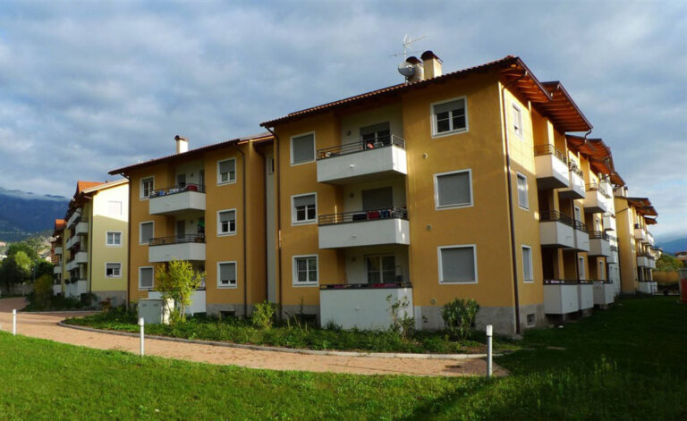 Architettura Moderna e Spazi Verdi: I Nostri Serramenti per Case ITEA a Riva del Garda (TN) Ampia veduta di una serie di edifici residenziali a più piani, probabilmente parte del complesso Case ITEA a Riva del Garda (TN), disposti lungo una curva. Gli edifici presentano facciate di diversi colori, prevalentemente giallo e beige, con balconi dotati di parapetti bianchi e grigi. Numerose finestre con infissi bianchi e avvolgibili grigi sono visibili su tutte le facciate. In primo piano, un'ampia area di prato verde ben curato e un percorso pedonale curvo. Sullo sfondo, si intravedono le montagne sotto un cielo nuvoloso ma luminoso.