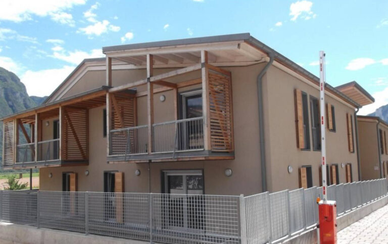 Architettura Moderna con Dettagli in Legno: Serramenti per Case ITEA a Rovere della Luna (TN) Immagine esterna di un edificio residenziale moderno, probabilmente parte del complesso Case ITEA a Rovere della Luna (TN), con facciate color tortora. L'edificio presenta ampi balconi con parapetti in metallo grigio e sovrastanti strutture in legno a doghe orizzontali che fungono da frangisole o elementi decorativi. Sono visibili finestre con infissi scuri e alcune persiane in legno a libro. In primo piano, una recinzione in rete metallica grigia e una sbarra d'ingresso automatica rossa e bianca. Sullo sfondo, si intravedono le montagne e un cielo azzurro con nuvole sparse.