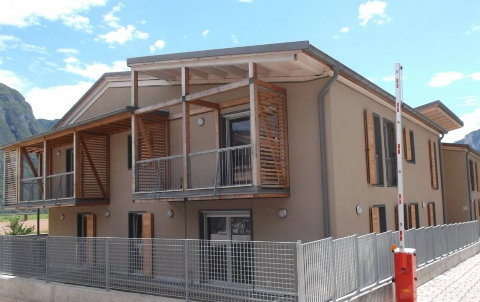 Architettura Moderna con Dettagli in Legno: Serramenti per Case ITEA a Rovere della Luna (TN) Immagine esterna di un edificio residenziale moderno, probabilmente parte del complesso Case ITEA a Rovere della Luna (TN), con facciate color tortora. L'edificio presenta ampi balconi con parapetti in metallo grigio e sovrastanti strutture in legno a doghe orizzontali che fungono da frangisole o elementi decorativi. Sono visibili finestre con infissi scuri e alcune persiane in legno a libro. In primo piano, una recinzione in rete metallica grigia e una sbarra d'ingresso automatica rossa e bianca. Sullo sfondo, si intravedono le montagne e un cielo azzurro con nuvole sparse.