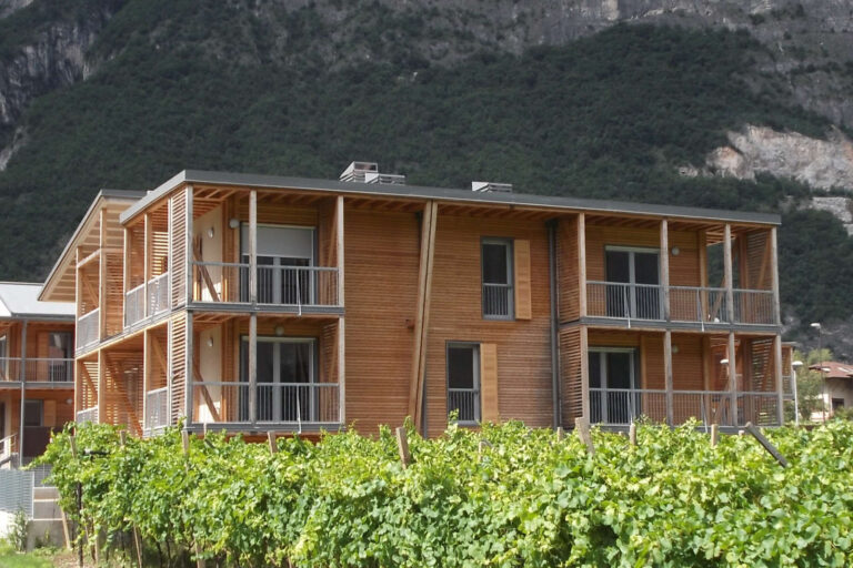 Architettura Residenziale Integrata nel Paesaggio: I Nostri Serramenti per Case ITEA a Rovere della Luna (TN) Immagine esterna di un edificio residenziale moderno a più piani, probabilmente parte del complesso Case ITEA a Rovere della Luna (TN), con facciate rivestite in legno chiaro e ampi balconi. I balconi sono dotati di parapetti a ringhiera orizzontale in metallo grigio e colonne portanti in legno che si estendono fino al tetto. Si notano numerose finestre con infissi scuri e alcune persiane in legno a libro. L'edificio è immerso in un paesaggio verde, con un vigneto in primo piano e montagne rocciose sullo sfondo. Il cielo è azzurro e limpido.