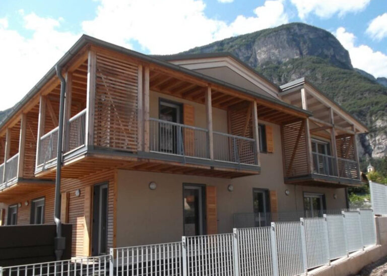 Finestre e Chiusure Innovative per Complesso Residenziale ITEA a Rovere della Luna (TN) Immagine esterna di un edificio residenziale moderno, probabilmente parte del complesso Case ITEA a Rovere della Luna (TN), con facciate color tortora e dettagli in legno chiaro. L'edificio presenta ampi balconi con parapetti in metallo grigio e sovrastanti strutture in legno a doghe orizzontali che fungono da frangisole o elementi decorativi. Sono visibili finestre con infissi scuri e alcune persiane in legno a libro. In primo piano, una recinzione in metallo grigio. Sullo sfondo, si stagliano le imponenti montagne, tipiche del paesaggio trentino, sotto un cielo azzurro con nuvole sparse.