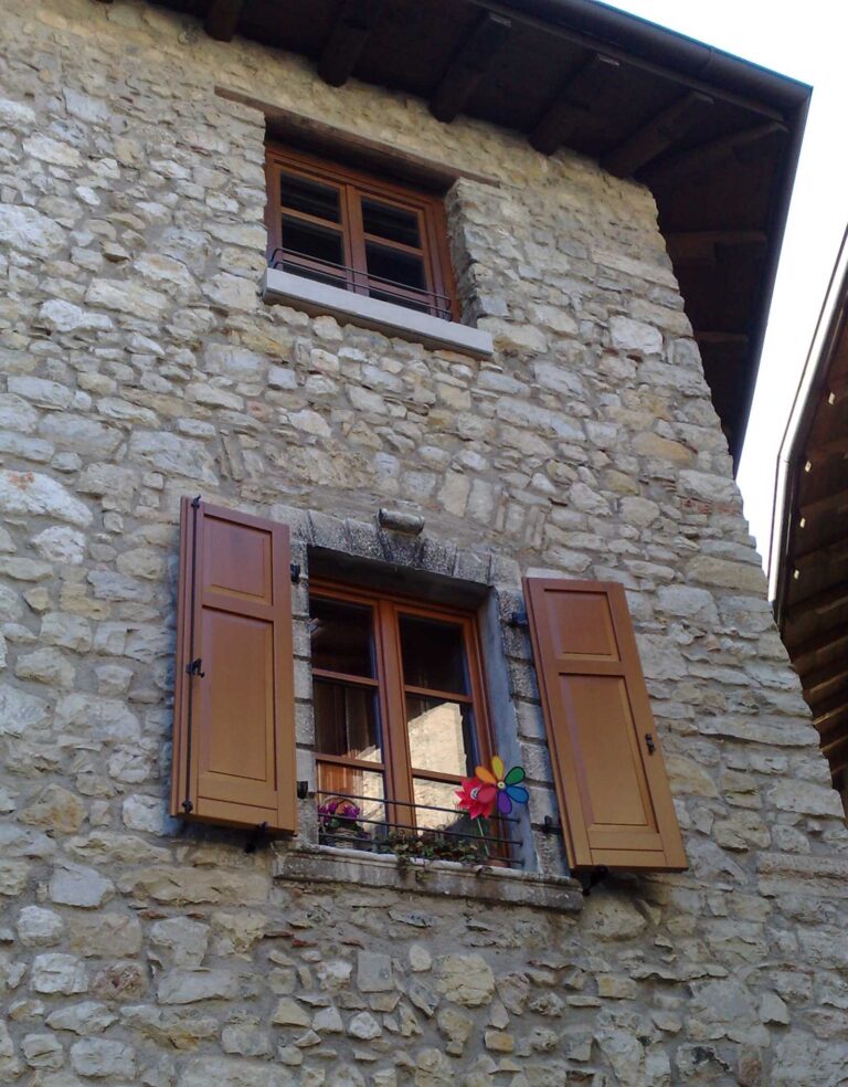 Restauro Finestre e Chiusure per Edifici Storici a Canale di Tenno: Immobiliare Omega (TN) Veduta ravvicinata di una porzione di facciata di un edificio in pietra, probabilmente parte di Immobiliare Omega a Canale di Tenno (TN). La parete è rivestita in pietra a vista e presenta due finestre con infissi in legno marrone. La finestra inferiore è dotata di persiane in legno marrone aperte e ha una fioriera con un mulinello a vento colorato sul davanzale. La finestra superiore è più piccola e senza persiane. Un tetto spiovente scuro è visibile nella parte superiore dell'immagine.