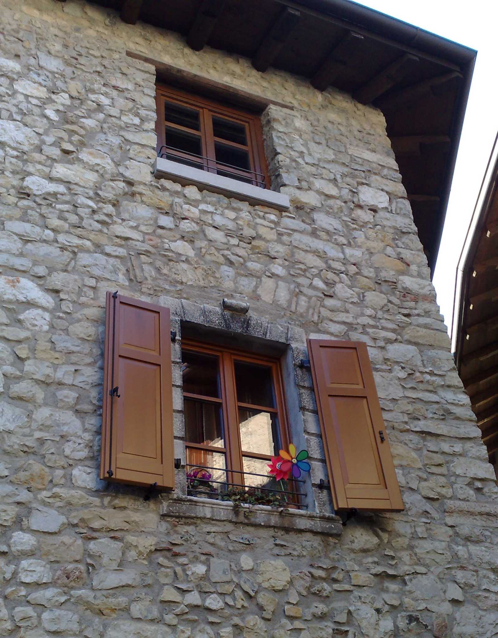 Restauro Finestre e Chiusure per Edifici Storici a Canale di Tenno: Immobiliare Omega (TN) Veduta ravvicinata di una porzione di facciata di un edificio in pietra, probabilmente parte di Immobiliare Omega a Canale di Tenno (TN). La parete è rivestita in pietra a vista e presenta due finestre con infissi in legno marrone. La finestra inferiore è dotata di persiane in legno marrone aperte e ha una fioriera con un mulinello a vento colorato sul davanzale. La finestra superiore è più piccola e senza persiane. Un tetto spiovente scuro è visibile nella parte superiore dell'immagine.