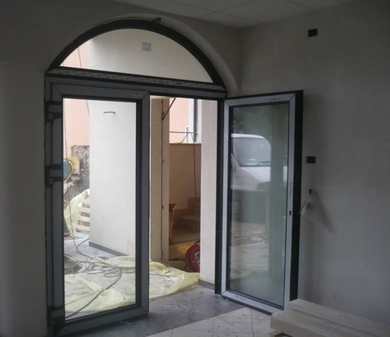 Porte e Chiusure per Edifici a Canale di Tenno: Interni ed Esterni per Immobiliare Omega (TN) Immagine interna di un ingresso, presso Immobiliare Omega a Canale di Tenno (TN), con un'ampia porta a doppio battente in vetro e cornice scura che si apre verso l'esterno. Sopra la porta, un'apertura ad arco vetrata permette il passaggio di ulteriore luce. Il pavimento è di cemento con alcune lastre gialle di protezione, e l'ambiente appare in fase di ristrutturazione. Sullo sfondo, si intravede una scala e un'area esterna.