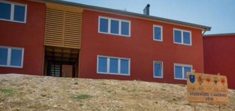 Edilizia Pubblica: Serramenti e Facciate Ventilate per Studentato Camerino (MC) Immagine esterna di un moderno edificio a più piani con facciate di colore rosso vivace e finestre rettangolari con infissi bianchi. Al centro dell'edificio si nota un grande rivestimento verticale in listelli di legno chiaro, che cela un'apertura o un ingresso. In primo piano a destra, c'è un cartello in legno con la scritta "STUDENTATO CAMERINO 2018" e alcuni stemmi o loghi, indicando l'anno di realizzazione. L'edificio è situato su un terreno in pendenza con terra e poca vegetazione, suggerendo una fase post-costruzione o riqualificazione. Il cielo è azzurro e sereno, e in lontananza si intravedono altri edifici.