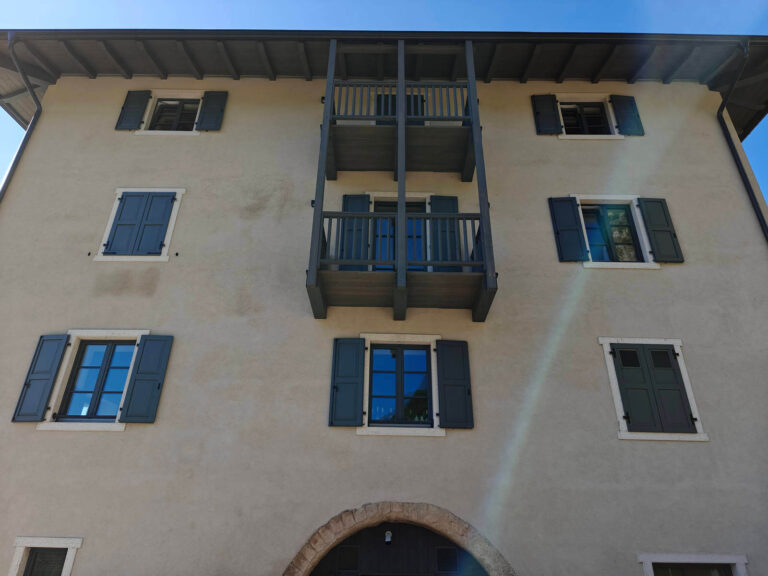 Design Rustico Moderno e Infissi di Qualità: I Nostri Serramenti per la Garda Stube di Riva del Garda (TN) Veduta frontale di una facciata dell'edificio dove al piano terra c'è il ristorante Garda Stube a Riva del Garda (TN), caratterizzata da finestre con scuri grigi a battente e un balcone centrale in legno a due livelli. La facciata è di colore chiaro, con un'apertura ad arco in pietra alla base. Il tetto è sporgente con travi a vista. Si notano ombre proiettate dal sole sulla facciata, indicando una buona esposizione alla luce.